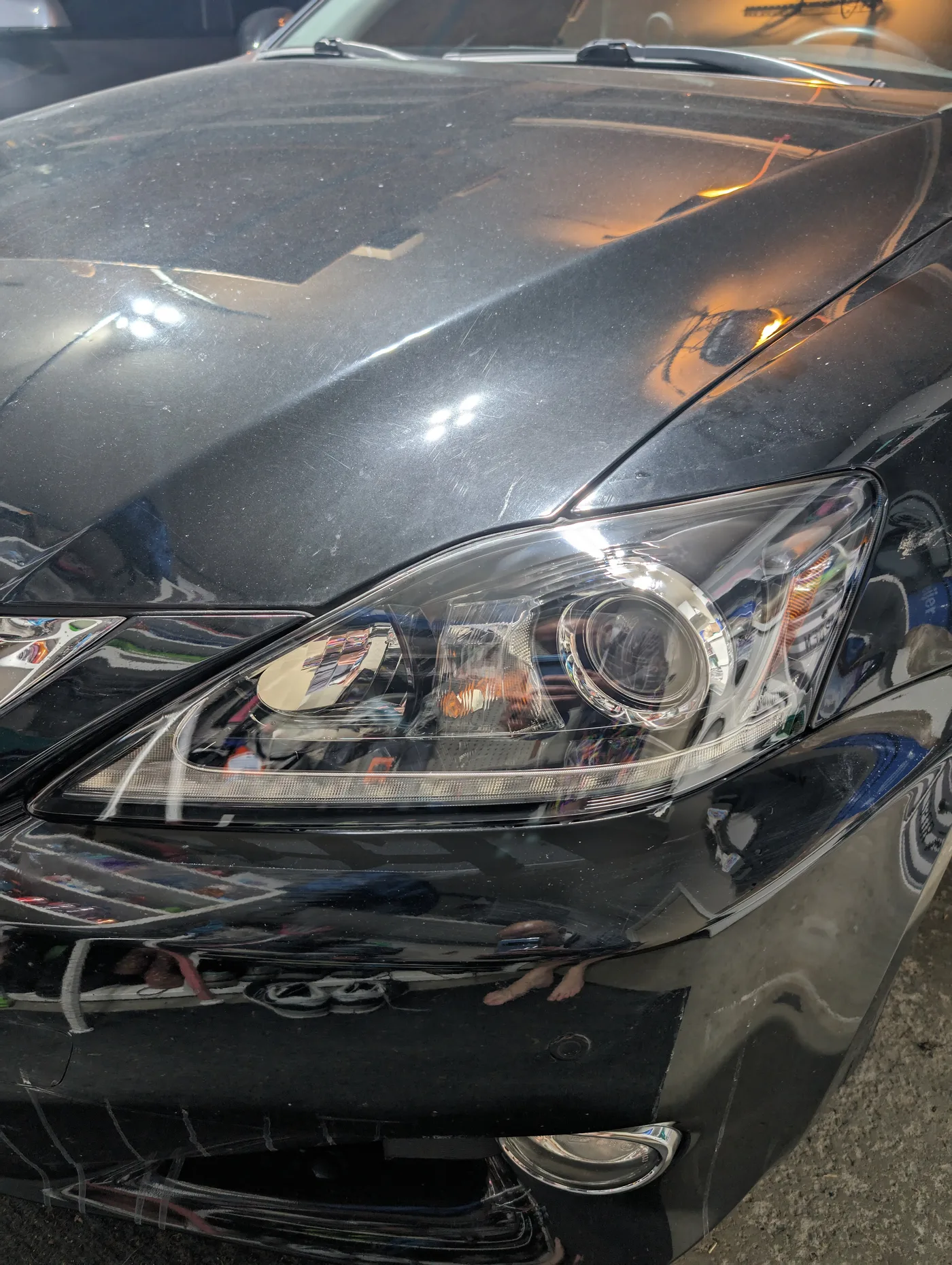 Front corner of a black Lexus IS under shop lighting, showing fine scratches and swirl marks across the hood